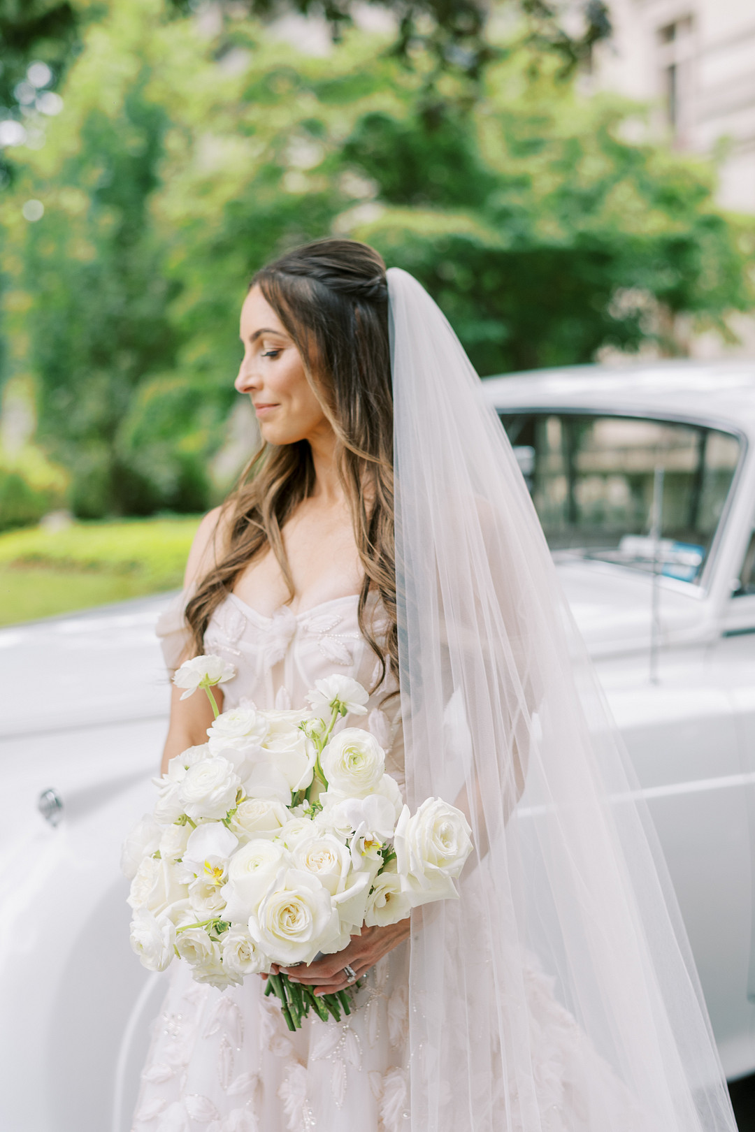 Bride holding a white bouquet beside a vintage white car