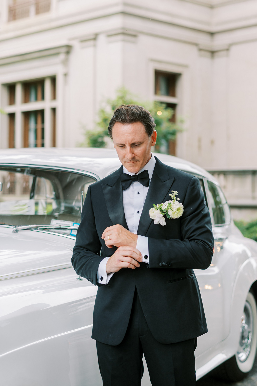 Groom in a black tuxedo adjusting his cufflinks beside a vintage car