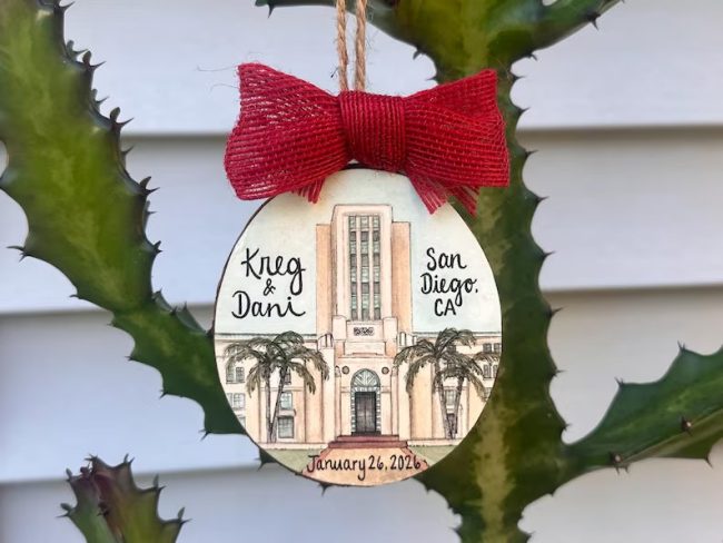 Wood plank ornament with a watercolor painting of a venue building, sample names and a date, topped with a red bow