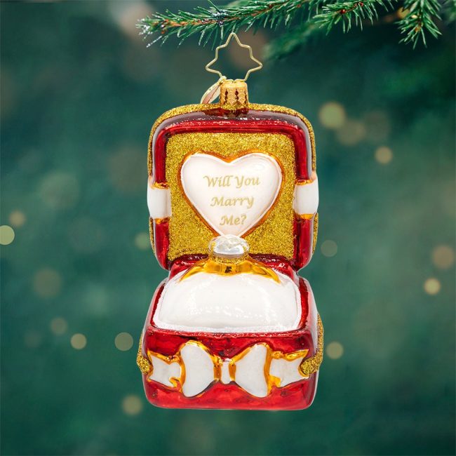 Christopher Radko glass ornament shaped like an open red and gold ring box with a Will You Marry Me heart inside, hanging on a tree