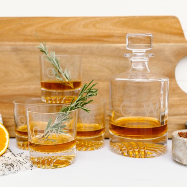 Glass whiskey decanter and four rocks glasses etched with a monogram, filled with whiskey and garnished with rosemary on a wooden board