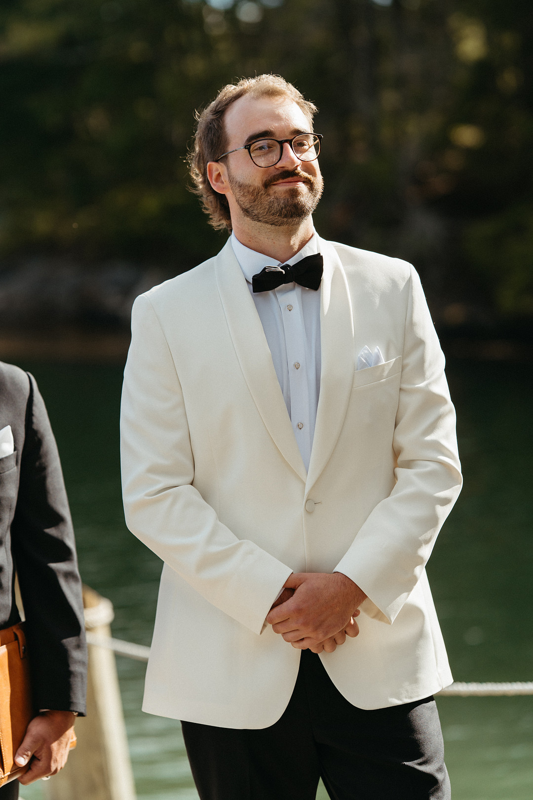 Groom in an off-white tuxedo jacket and black bow tie by the river