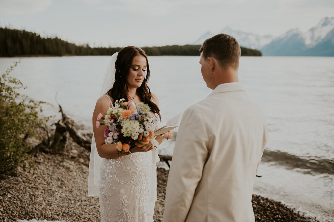 Bride holding a colorful bouquet as the groom reads his vows by the lake
