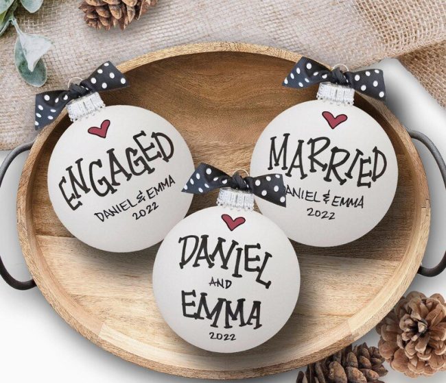 Three white glass ornaments hand-lettered Engaged, sample names and Married, with polka dot bows, on a wood tray