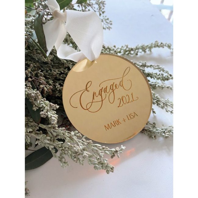Round wood ornament engraved with Engaged in script and a year, hanging from a white ribbon