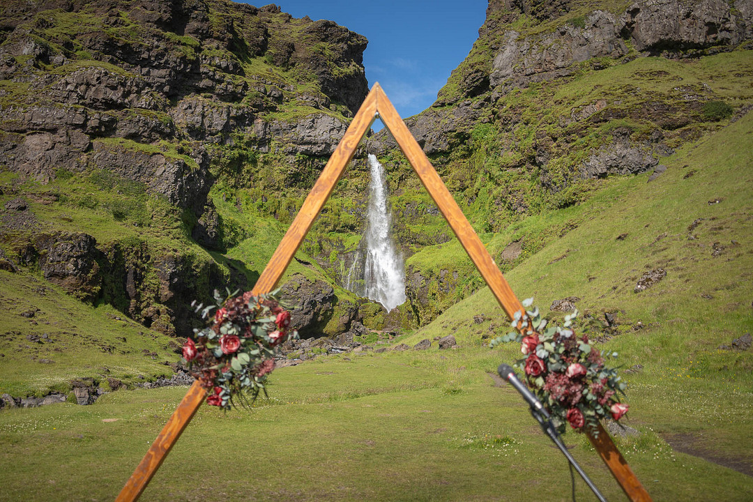 Triangular wooden ceremony arch with red and pink flowers in front of a waterfall