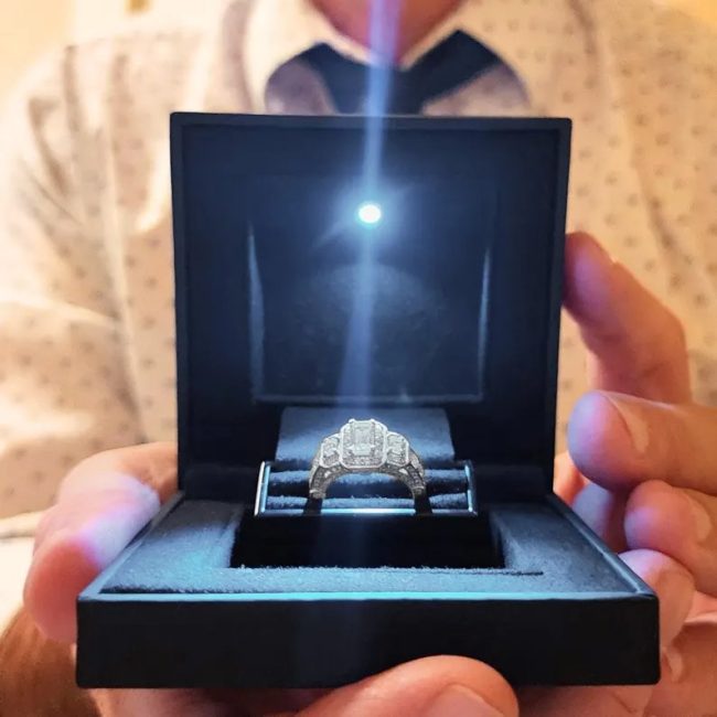 Hands holding an open LittleOlivers black ring box lit from inside, with the ring raised on its center holder
