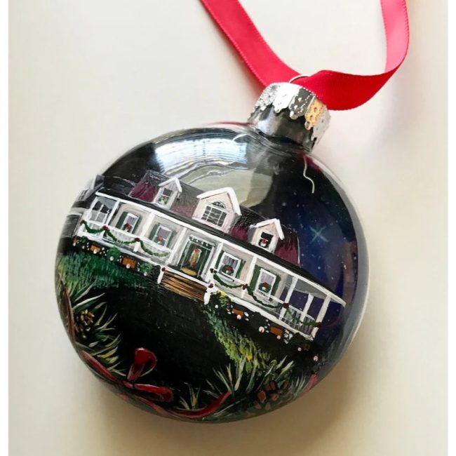 Dark glass disc ornament hand-painted with a white two-story house and trees, hanging from a red ribbon