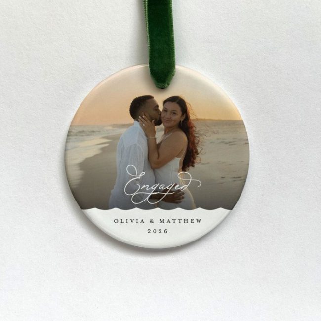 Minted round ceramic ornament with a couple's beach photo, Engaged in script and sample names, on a green ribbon