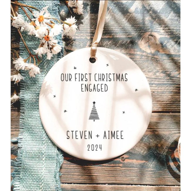 Round white ceramic ornament reading Our First Christmas Engaged with a small tree and sample names, on a wooden table