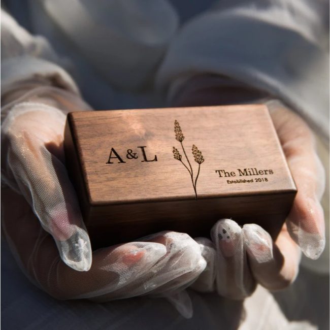 Gloved hands holding a MUUJEE walnut ring box engraved with the initials A and L