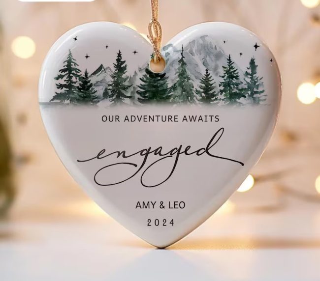 Heart-shaped white ceramic ornament with a watercolor mountain and pine tree scene and the word engaged in script