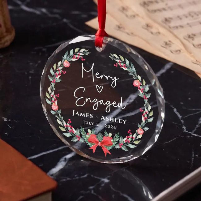 Clear faceted glass ornament printed with a holly wreath and Merry Engaged with sample names, on dark marble