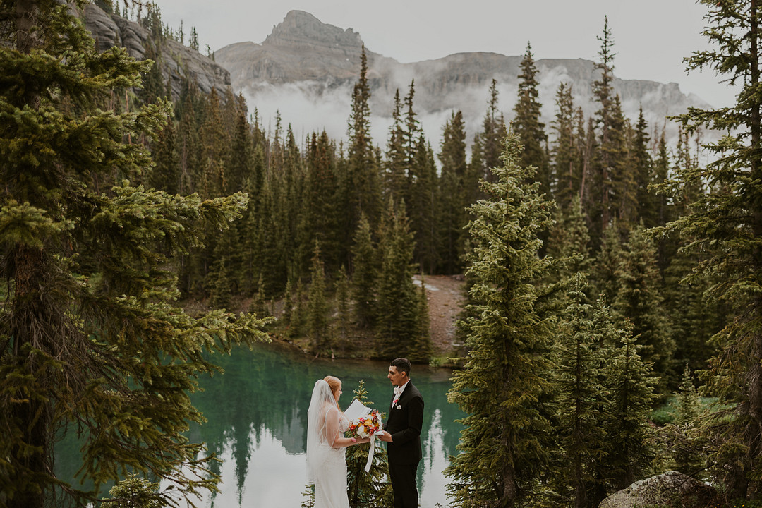 Wide view of the couple beside the lake surrounded by evergreens and mountains
