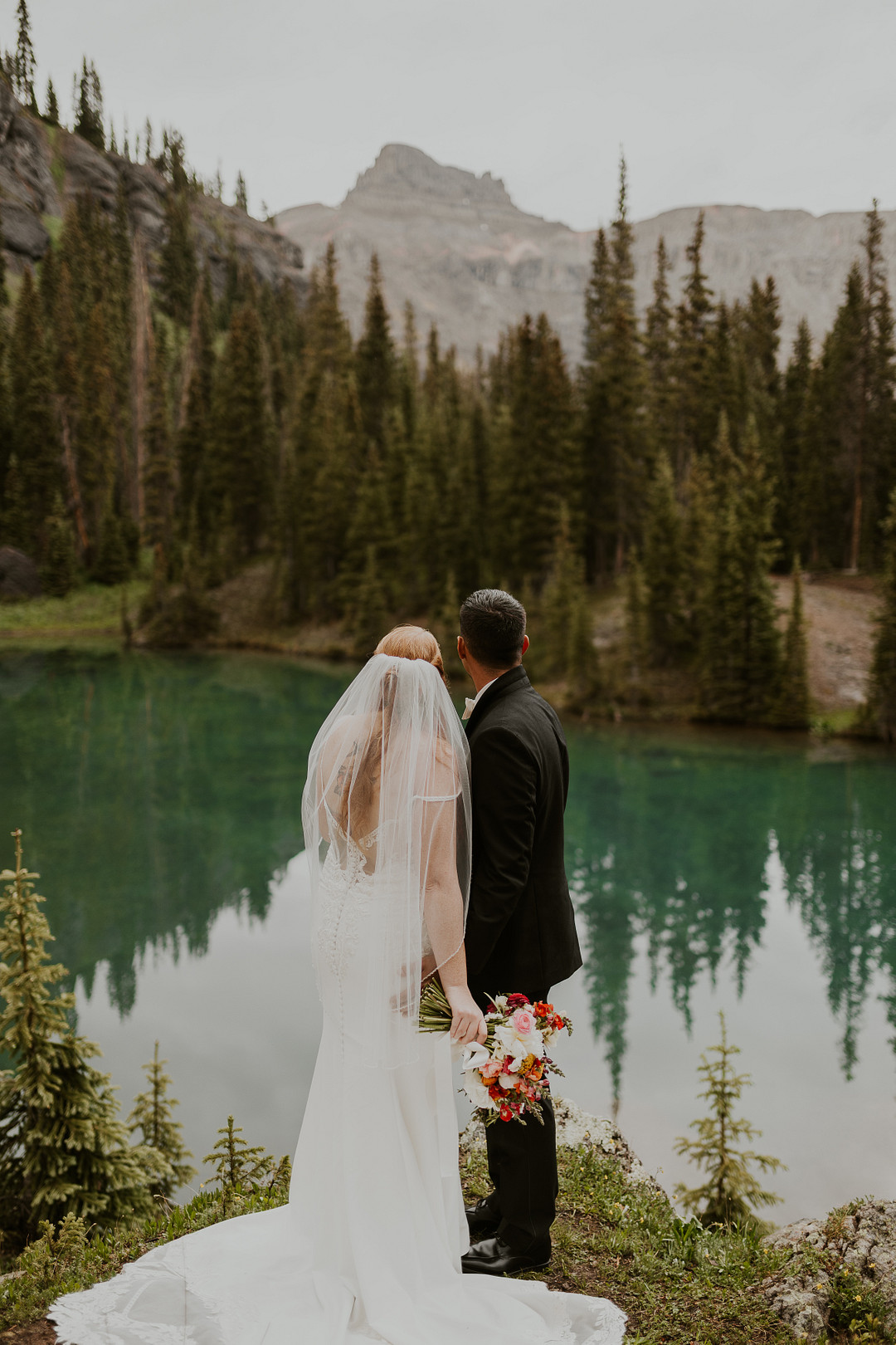 Bride and groom standing among evergreens at the turquoise lake