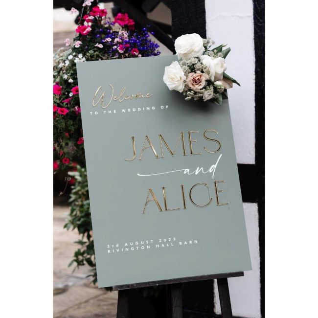 RoseSignageCompany sage acrylic welcome sign with mirrored names on an easel, topped with white flowers
