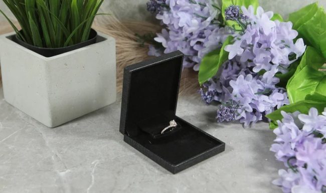 Soulmatebox slim gray ring box open to show a diamond ring, beside purple flowers