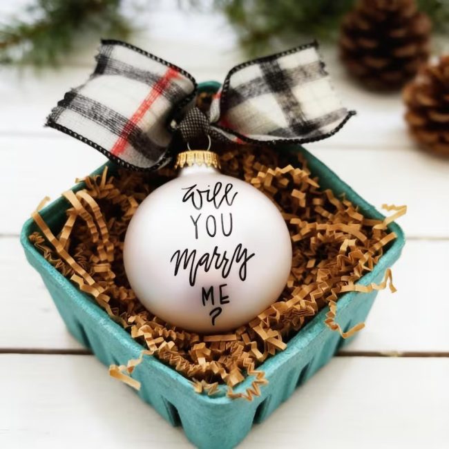 White glass ball ornament reading Will You Marry Me with a buffalo check ribbon, in a basket of paper shred