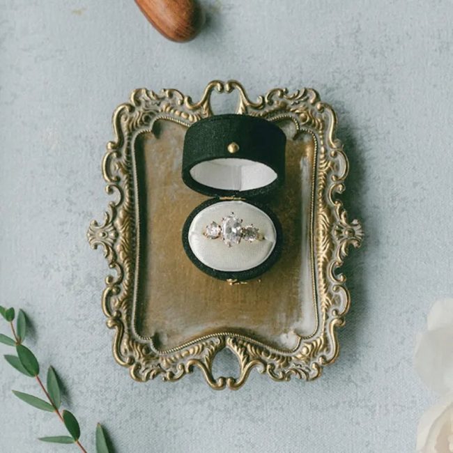 TheHeirloomWedding green linen and velvet oval ring box open on a gold tray, holding a diamond ring