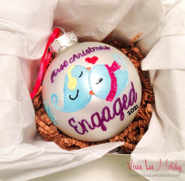 White glass ornament hand-painted with two blue lovebirds and First Christmas Engaged in pink glitter, in a gift box