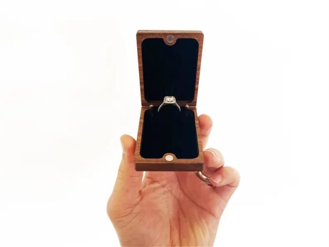 Hand holding an open Wanderweg slim walnut flip-up ring box with a solitaire ring inside