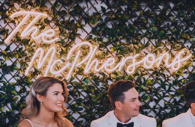 ZanvisNeon warm white neon sign spelling The McPhersons on a greenery wall above a smiling bride and groom